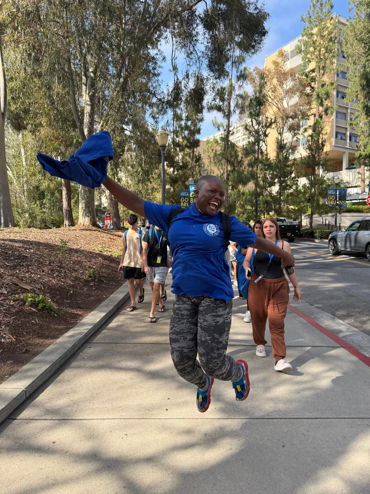 An ELC counselor in a blue staff shirt jumping in the air on a walkway on the UCLA campus